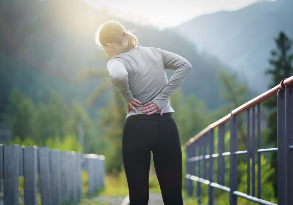A woman holds her lower back in pain outdoors, with mountains in the background.